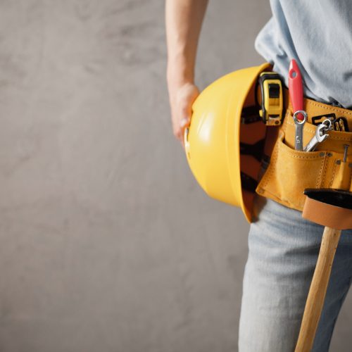 Worker man and tool belt holding construction helmet near wall. Male hand and tools